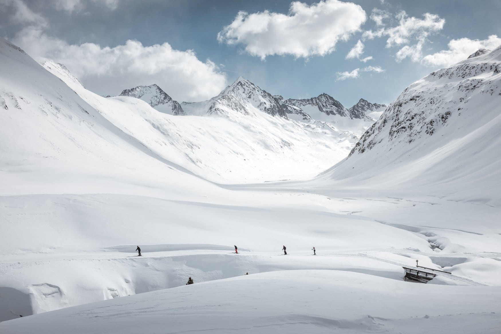Vier Skifahrer ziehen hintereinander über einen Weg durch ein tief verschneites Hochtal bei der Schönwieshütte, im Hintergrund ragen schneebedeckte Gipfel in den bewölkten Himmel.