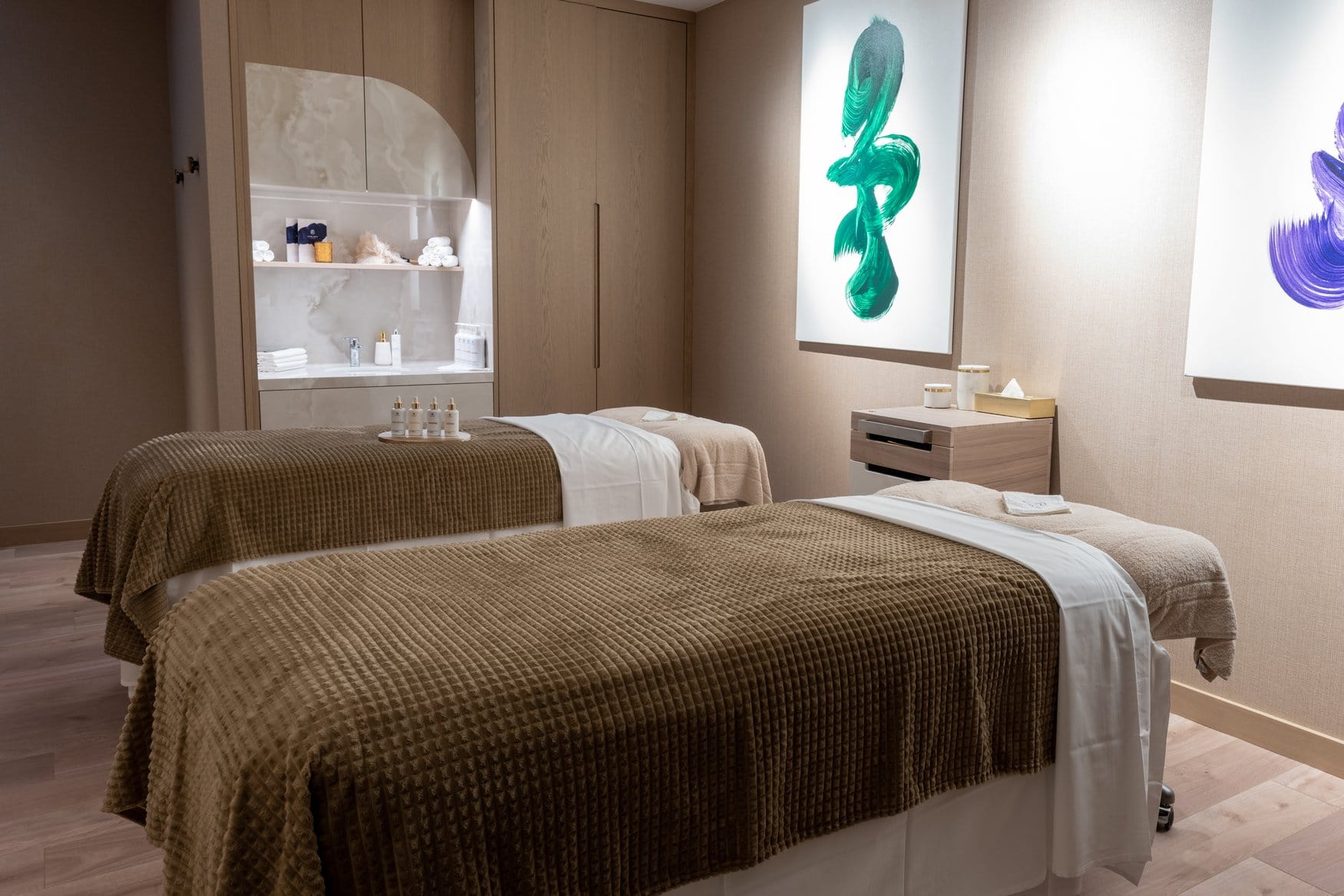 Behandlungsraum im Spa mit zwei Massageliegen unter beigen Waffeldecken. An der Wand hängen hinterleuchtete Bilder mit grünen und violetten Pinselschwüngen, links eine Nische mit Waschtisch und gestapelten Handtüchern.