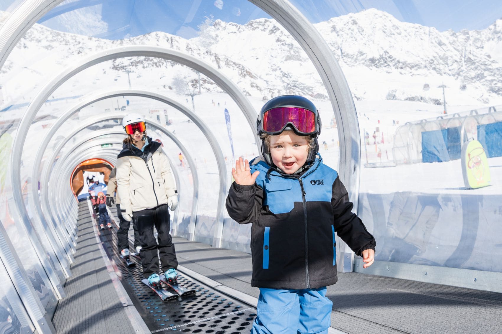 Kind mit Skihelm und Skibrille winkt auf einem überdachten Förderband im Kinderland am Stubaier Gletscher in Tirol.