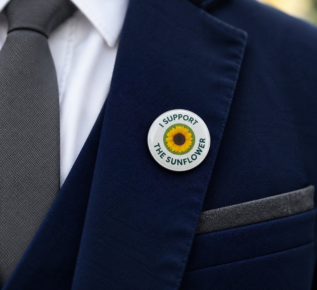 Runder weißer Anstecker mit Sonnenblume und der Aufschrift »I support the Sunflower« am Revers eines marineblauen Sakkos mit grauer Krawatte.