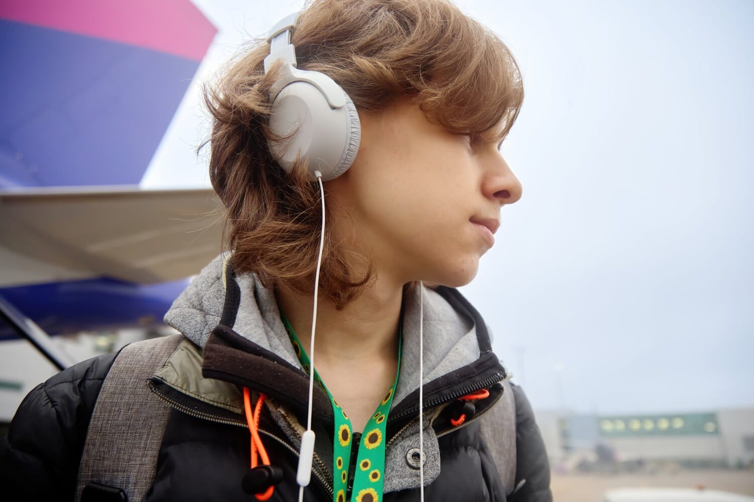 Junge Person mit weißen Kopfhörern und grünem Sunflower-Lanyard um den Hals steht auf dem Vorfeld vor einem Flugzeugheck und blickt zur Seite.
