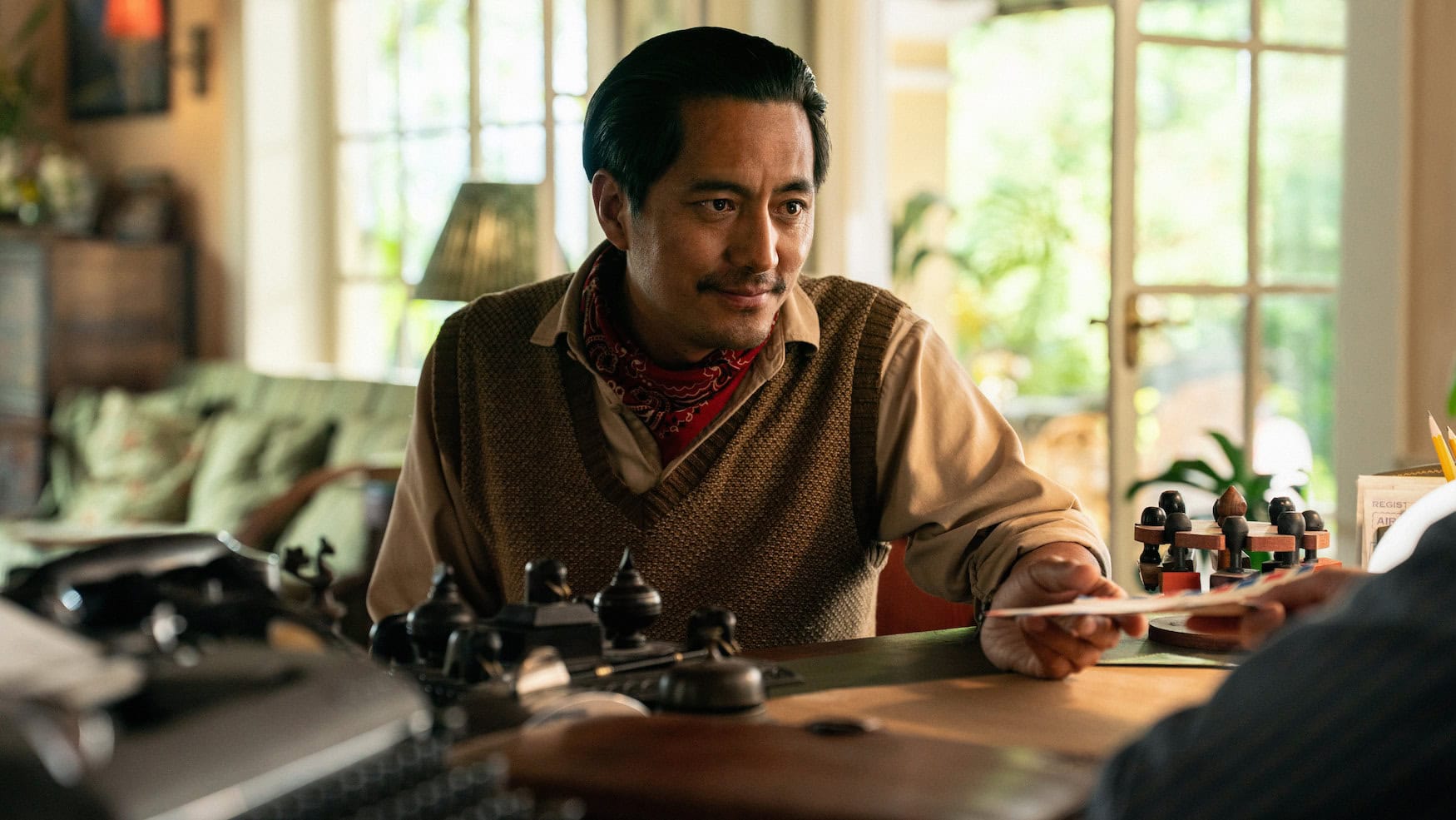 Ein Mann mit brauner Strickweste und rotem Halstuch sitzt an einem Schreibtisch und reicht einer anderen Person ein Dokument. Szene aus dem Apple-TV-Film »Tenzing«.