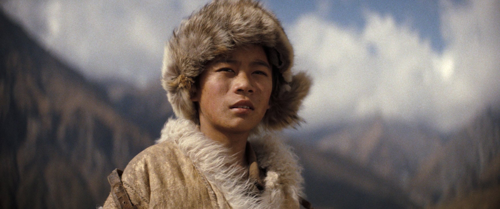 Ein junger Mann mit großer Fellmütze und fellgefütterter Kleidung steht vor einer Bergkulisse. Szene aus dem Apple-TV-Film »Tenzing«.