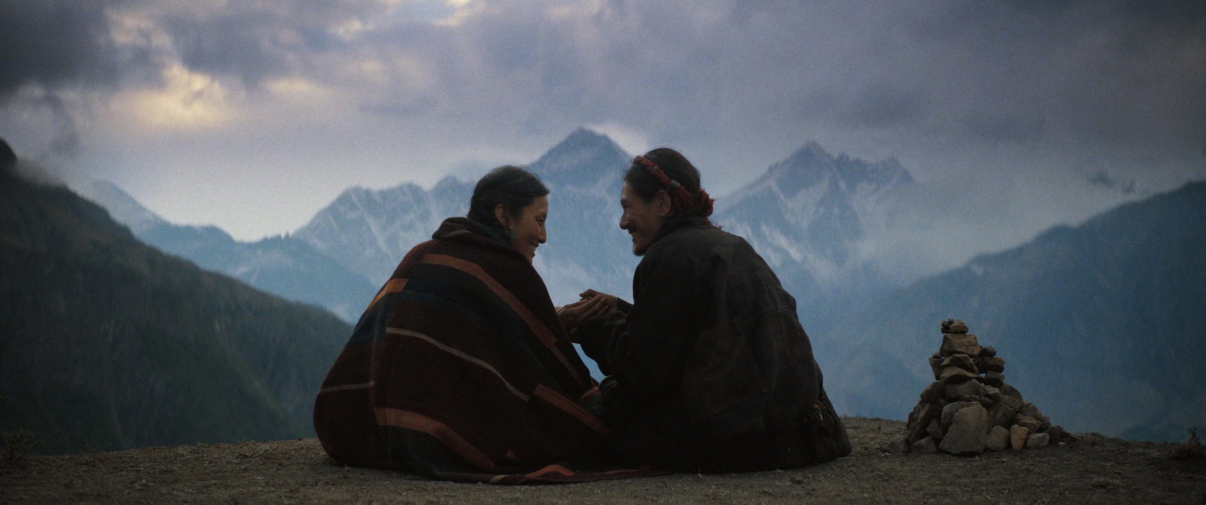 Zwei Personen sitzen eng beieinander auf einem Bergrücken und halten sich an den Händen. Im Hintergrund ragen schneebedeckte Gipfel unter einem wolkenverhangenen Himmel auf. Szene aus dem Apple-TV-Film »Tenzing«.