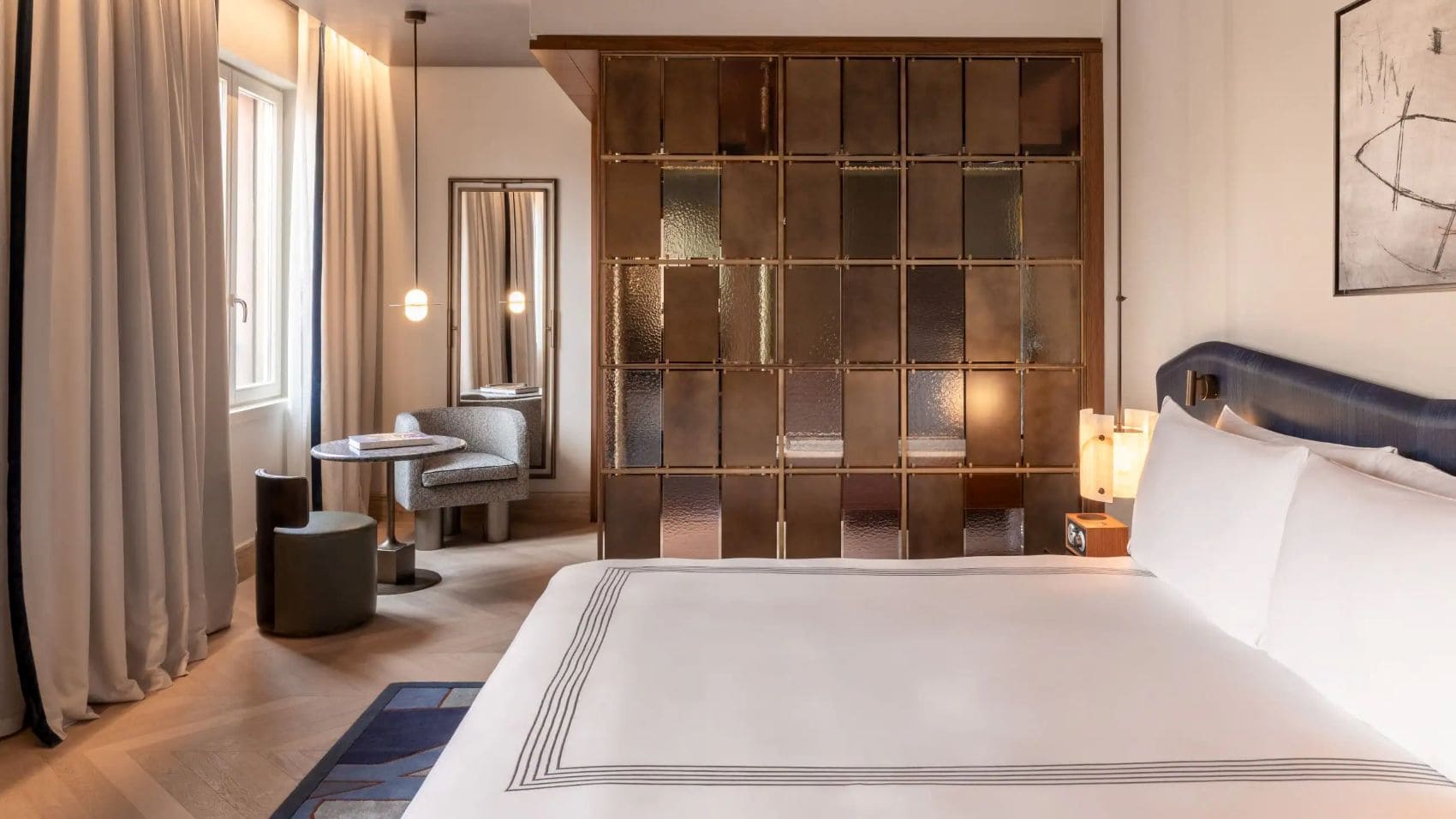 Deluxe-Doppelzimmer im Thompson Rome mit großem Bett, blauem Kopfteil, gläserner Raumtrennwand und Sitzecke am Fenster.