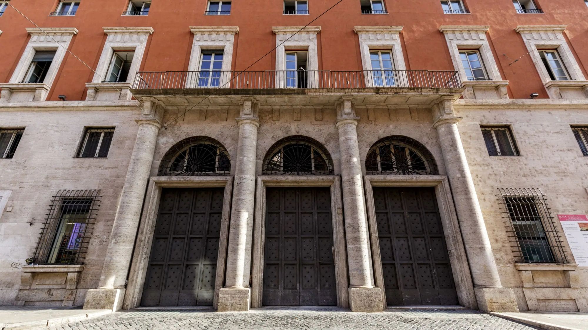 Historische Fassade des Thompson Rome in der Via delle Botteghe Oscure mit drei großen Eingangstoren, Steinsäulen und rot verputzten Obergeschossen.