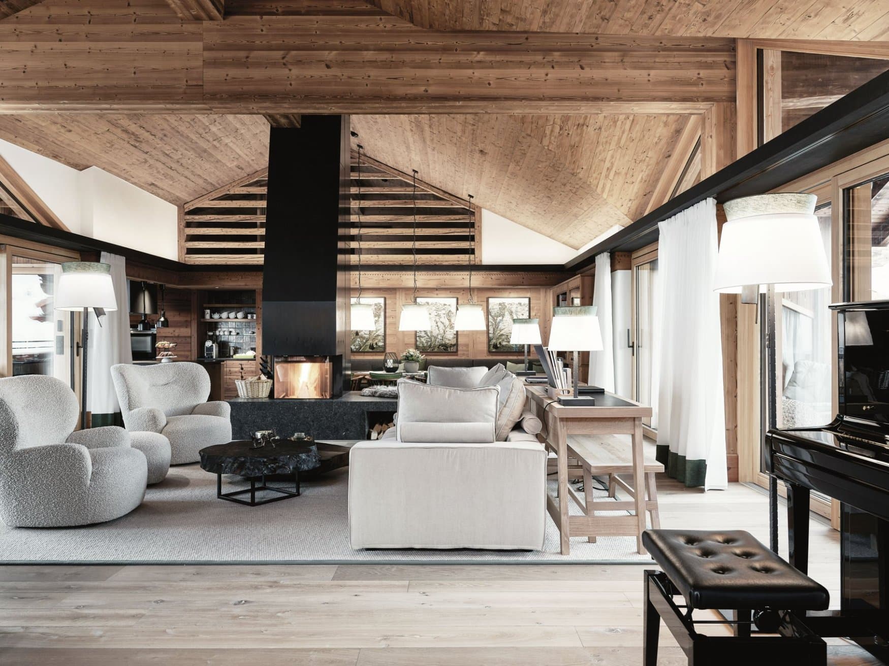 Ein offener Wohnraum unter einer Altholzdecke mit schwarzem Kaminblock in der Mitte, in dem ein Feuer brennt. Links stehen helle Bouclé-Sessel um einen Couchtisch aus einer Baumscheibe, rechts ein beigefarbenes Sofa vor einem Konsolentisch, am Bildrand ein schwarzer Flügel. Im Hintergrund die offene Küche.