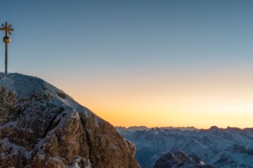 Goldenes Gipfelkreuz auf der verschneiten Zugspitze mit Alpenpanorama im warmen Licht.