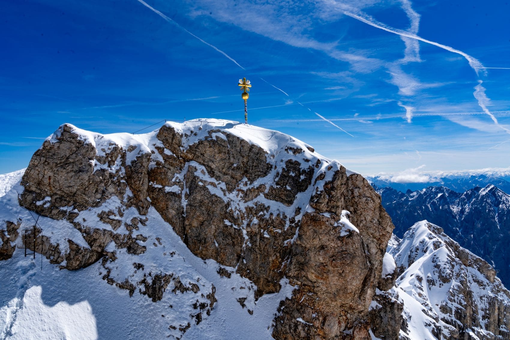 Goldenes Gipfelkreuz auf dem verschneiten Felsgipfel der Zugspitze vor blauem Himmel.