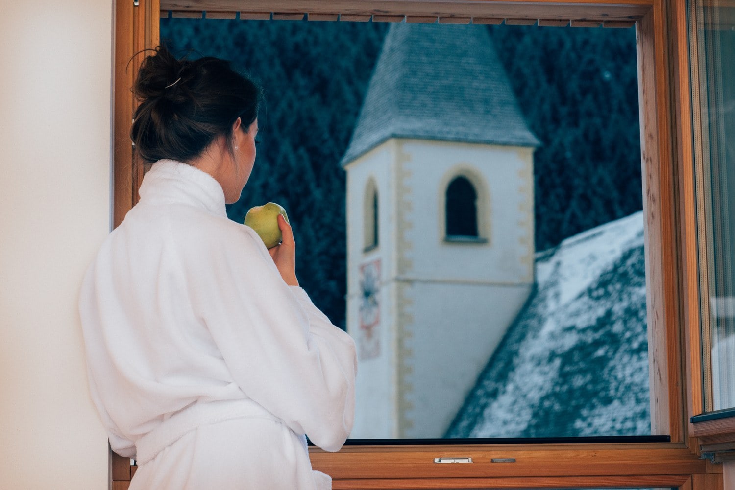 Frau im Bademantel blickt aus dem Fenster auf den verschneiten Kirchturm